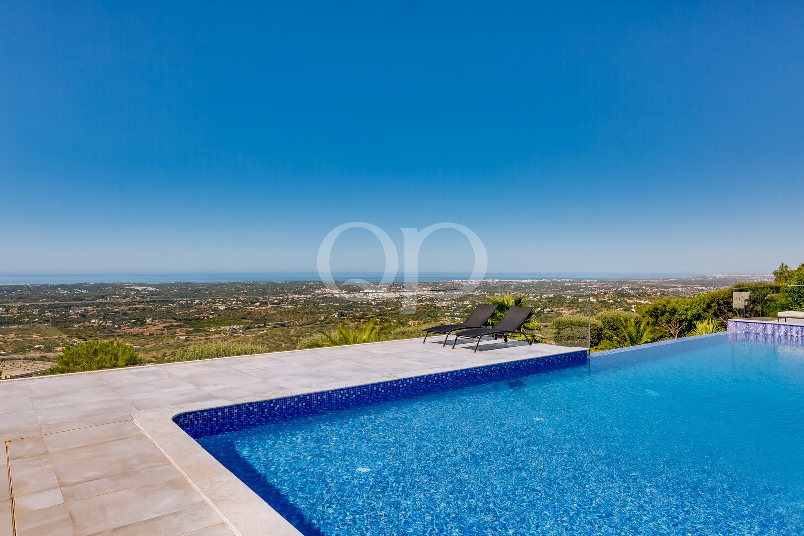 Infinity pool with blue water and two black lounge chairs on a stone deck overlooking a distant landscape and sea horizon under a clear blue sky