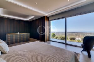 Modern bedroom with a large glass wall and balcony view of the landscape; bed in foreground and a striped dresser against a dark wood paneled wall.