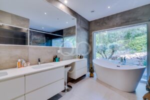 Modern bathroom with a white double vanity, large wall mirror, and a freestanding oval tub by a big window overlooking greenery.