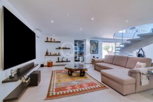 Modern living room with a large wall-mounted TV, beige sectional sofa, and a glass-topped round coffee table on a colorful rug.