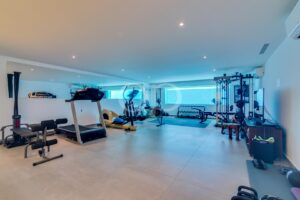 Home gym with treadmills, elliptical, weights, and a rack of barbells along a mirrored wall on the left; bright, airy space.