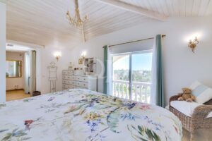 Bright bedroom with a white wooden vaulted ceiling, chandelier, and a sliding balcony door; floral duvet on the bed and a wicker chair with a teddy bear nearby.