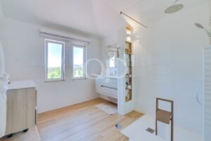 Bright modern bathroom with white tiles, two large windows overlooking greenery, and light wood flooring; features a floating vanity and open shelving.