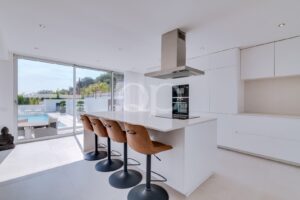 Helle, moderne weiße Küche mit einer zentralen Kochinsel, braunen Barhockern und Blick auf einen Außenpool durch große Glasschiebetüren.