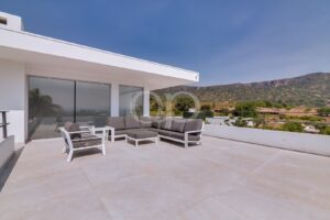 Eine weiße, moderne Terrasse mit einer grauen Sitzgruppe mit Polstern, von der aus man unter blauem Himmel auf die fernen Hügel blickt.