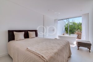Minimalistisches Schlafzimmer mit einem beigen, gesteppten Bett, einem dunklen Kopfteil und einer freistehenden weißen Badewanne neben großen Glasschiebetüren, die den Blick auf die grüne Umgebung im Freien freigeben.
