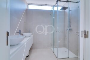 Modernes Badezimmer mit zwei weißen Aufsatzwaschbecken auf einem weißen Waschtisch, verchromten Armaturen und einer Duschkabine aus Glas mit einem großen Regenduschkopf.