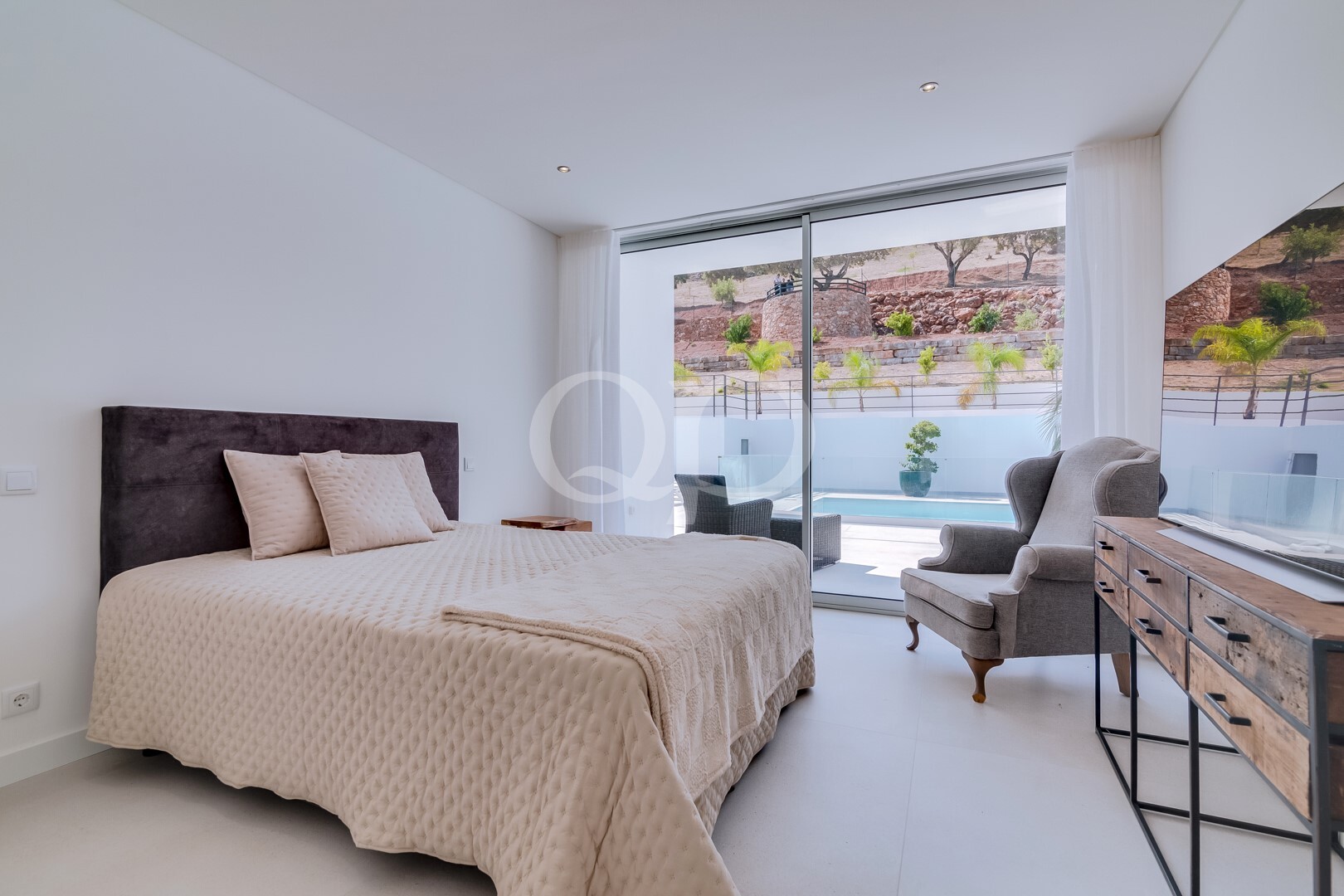 Schlafzimmer mit einem beigen Steppbett, zwei Kissen und einem dunklen Kopfteil; Glasschiebetüren führen hinaus zum Poolbereich.