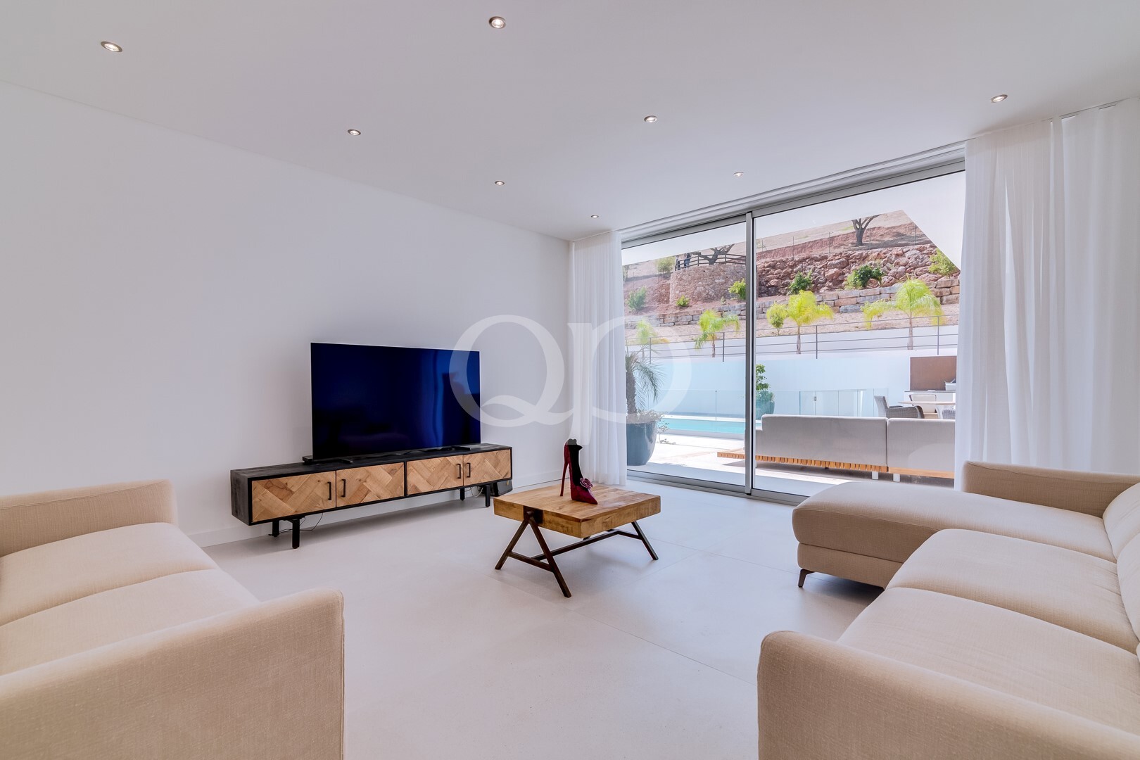 Helles, minimalistisch eingerichtetes Wohnzimmer mit beigen Sofas, einem TV-Schrank aus Holz und einem Flachbildfernseher; große Glastüren führen auf die Terrasse am Pool im Außenbereich.
