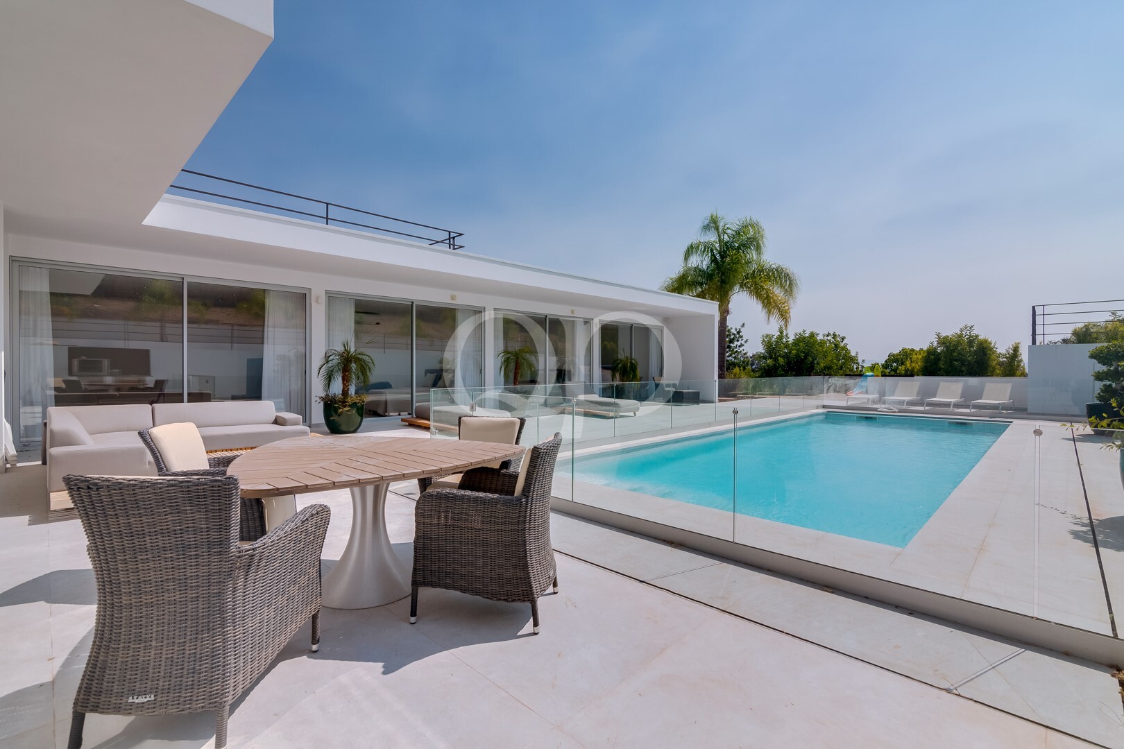 Moderne Terrasse mit Glasgeländer und Blick auf einen rechteckigen Pool, Liegestühle und eine Essgruppe aus Korbgeflecht in einem sonnigen Garten.
