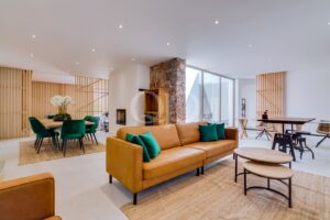 Offener Wohnbereich mit einem hellbraunen Ledersofa, blaugrünen Kissen, runden Couchtischen und einem Steinkamin in der Nähe großer Fenster.