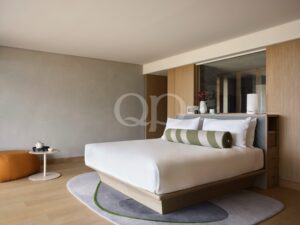 Modern hotel bedroom with white bedding, grey wall, and light wood headboard and cabinetry.