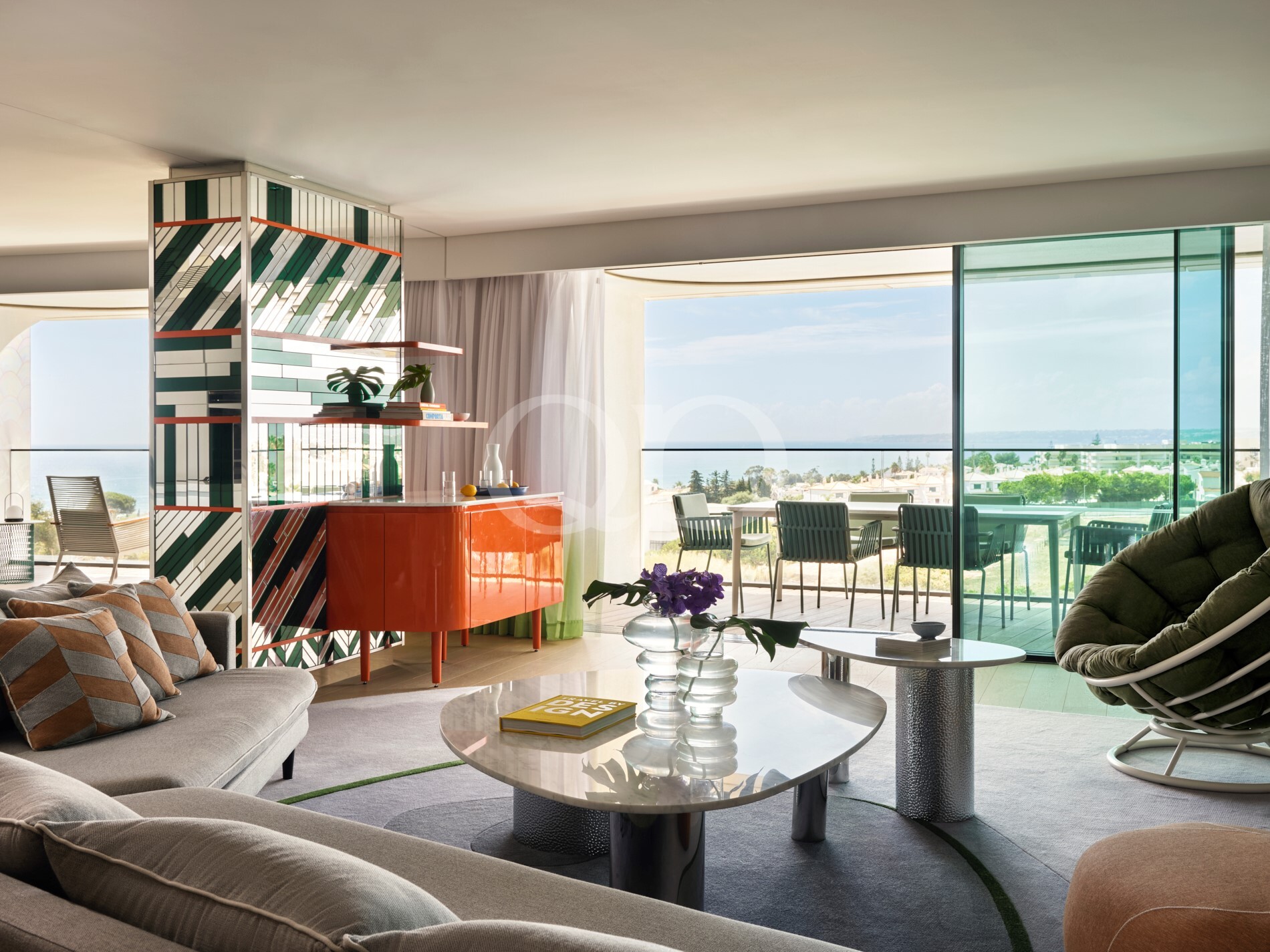 Modern living room with a geometric tiled divider, orange cabinet, and glass coffee tables, opening to a balcony with an ocean view.
