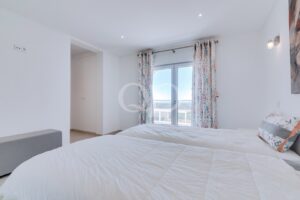 Quarto luminoso e minimalista, com duas camas com colchas brancas bem feitas, uma almofada decorativa, um banco à esquerda e portas de varanda com cortinas florais que deixam entrar a luz do dia.