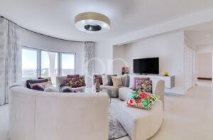Sala de estar moderna e luminosa, com um sofá seccional bege de linhas curvas, almofadas coloridas e uma mesa de centro de vidro em frente a uma televisão fixada na parede.