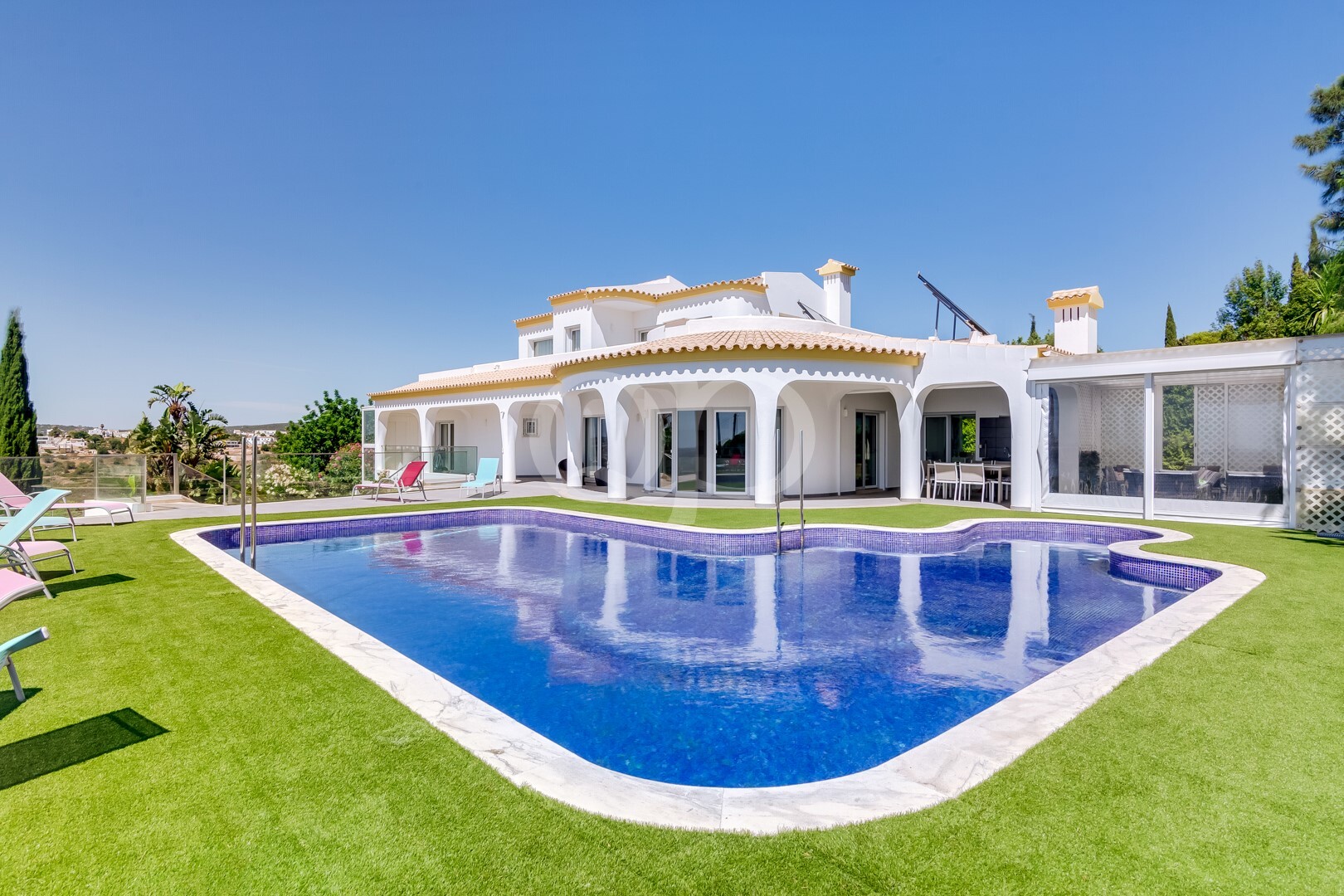 Eine weiße, moderne Villa mit geschwungenen Bögen, die an einem sonnigen Tag einen blauen Swimmingpool umgibt; Liegestühle stehen auf dem Rasen.