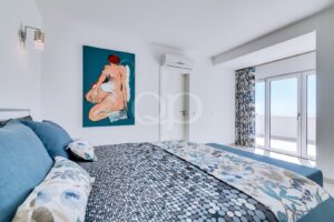 Quarto luminoso, ao estilo de hotel, com cabeceira azul, roupa de cama com padrão de bolinhas e um quadro em azul-petróleo representando uma figura nua na parede.