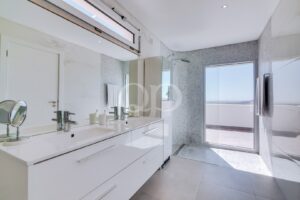 Casa de banho luminosa e moderna, com lavatório duplo, espelhos grandes e uma cabina de duche em vidro que dá acesso a uma varanda com vista para o mar.