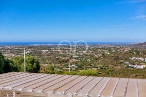 Vale costeiro panorâmico com colinas, casas dispersas e um oceano azul no horizonte; telhado metálico em primeiro plano.