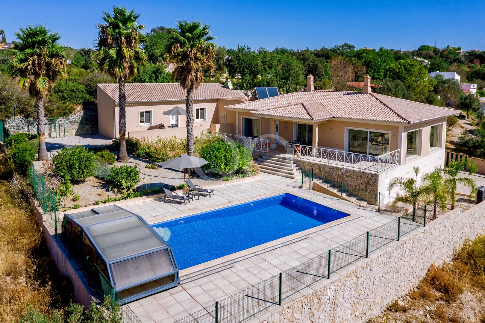 Une villa beige au toit en terre cuite, entourée de palmiers, donnant sur une piscine rectangulaire bleue et une terrasse carrelée.
