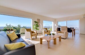 Sala de estar luminosa com vista para o litoral, com grandes janelas que revelam o céu azul e a vista para o mar; cadeiras bege, sofá creme e mesa de centro de madeira.