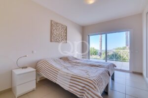 Quarto minimalista com um edredão bege às riscas, uma mesa de cabeceira branca e um candeeiro de leitura curvo junto à cama; à frente, as portas da varanda