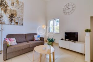 Sala de estar moderna com um sofá cinzento, almofadas coloridas e uma mesa de centro redonda de madeira, sob uma obra de arte abstrata na parede e paredes em tons neutros.
