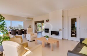 Sala de estar luminosa com poltronas de couro creme, uma mesa de centro de madeira e uma lareira, que dá acesso a uma zona de refeições e a uma varanda com vista para o exterior.