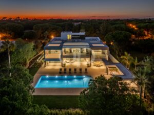 Luftaufnahme eines modernen mehrstöckigen Hauses mit beleuchtetem Pool und Dachterrasse in der Abenddämmerung, umgeben von Bäumen.