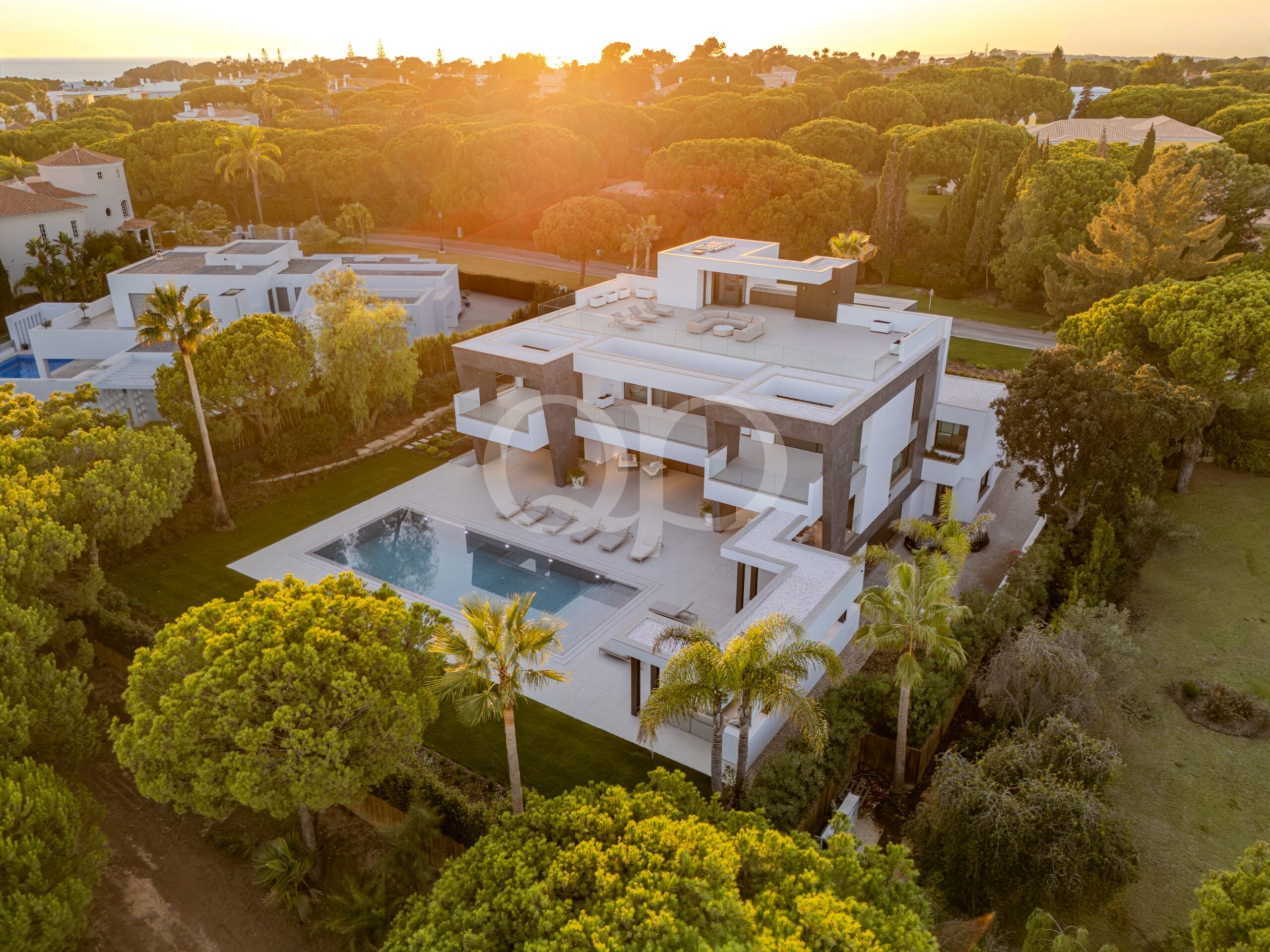 Vista aérea de uma villa moderna branca com uma grande piscina enterrada, rodeada por árvores frondosas ao pôr-do-sol.