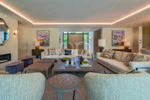 Helles, modernes Wohnzimmer mit beigen Ecksofas, abstrakter Wandkunst und einem zentralen Couchtisch aus Holz, der in warmes Licht aus Einbauleuchten getaucht ist.