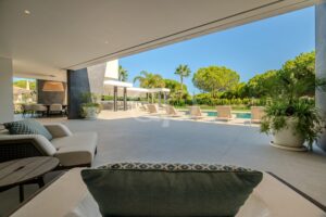 Lounge im Freien am Pool mit beigen Liegestühlen und einer schattigen Sitzecke unter einer überdachten Terrasse, darüber der blaue Himmel.