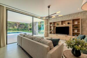 Modernes Wohnzimmer mit einem beigen Ecksofa, eingebauten Holzregalen und einem Flachbildfernseher, mit Blick auf große Glastüren, die zum Garten führen.