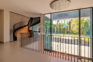Modernes Interieur: Eine geschwungene Treppe aus schwarzem Metall entlang einer hellen Wand, mit einer großen Glaswand, durch die man die grünen Bäume draußen sehen kann, und hängenden Pendelleuchten über dem Geländer.