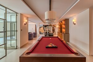 Luxuriöses Spielzimmer mit einem roten Billardtisch im Vordergrund und einem Loungebereich im Hintergrund mit warmer Beleuchtung.