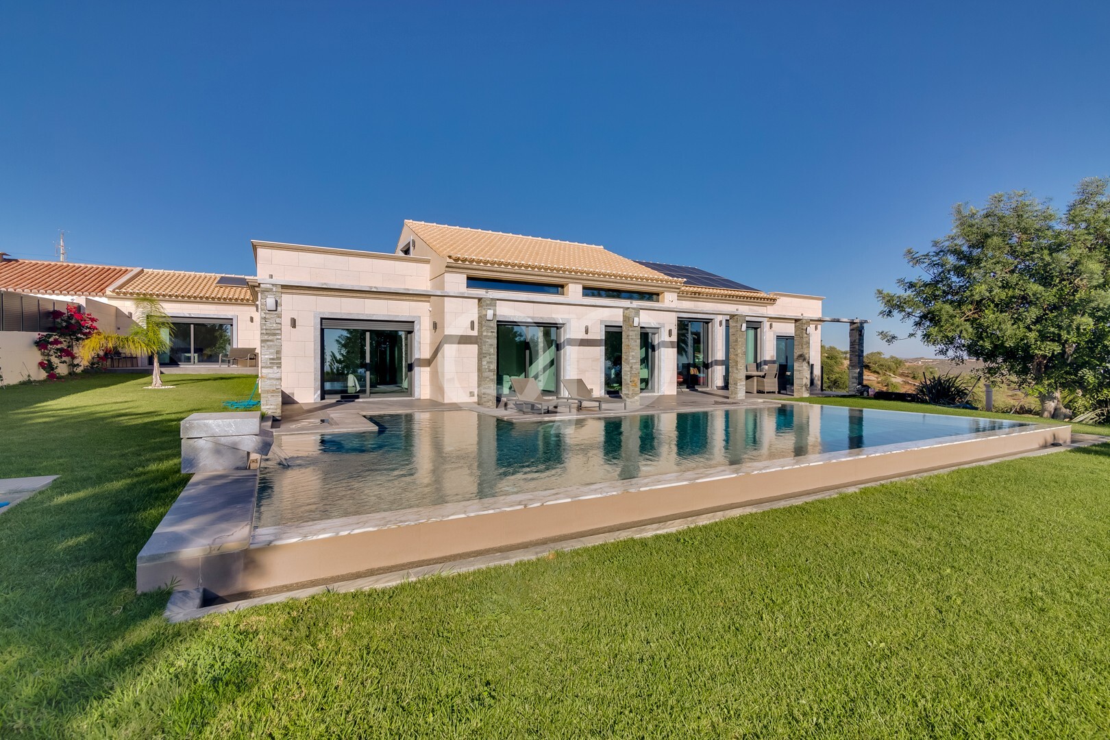 Villa moderne de plain-pied dotée de grandes baies vitrées, d'une façade en pierre beige et en tuiles, ainsi que d'une longue piscine rectangulaire entourée d'une pelouse verte et de chaises longues, sous un ciel d'un bleu limpide.