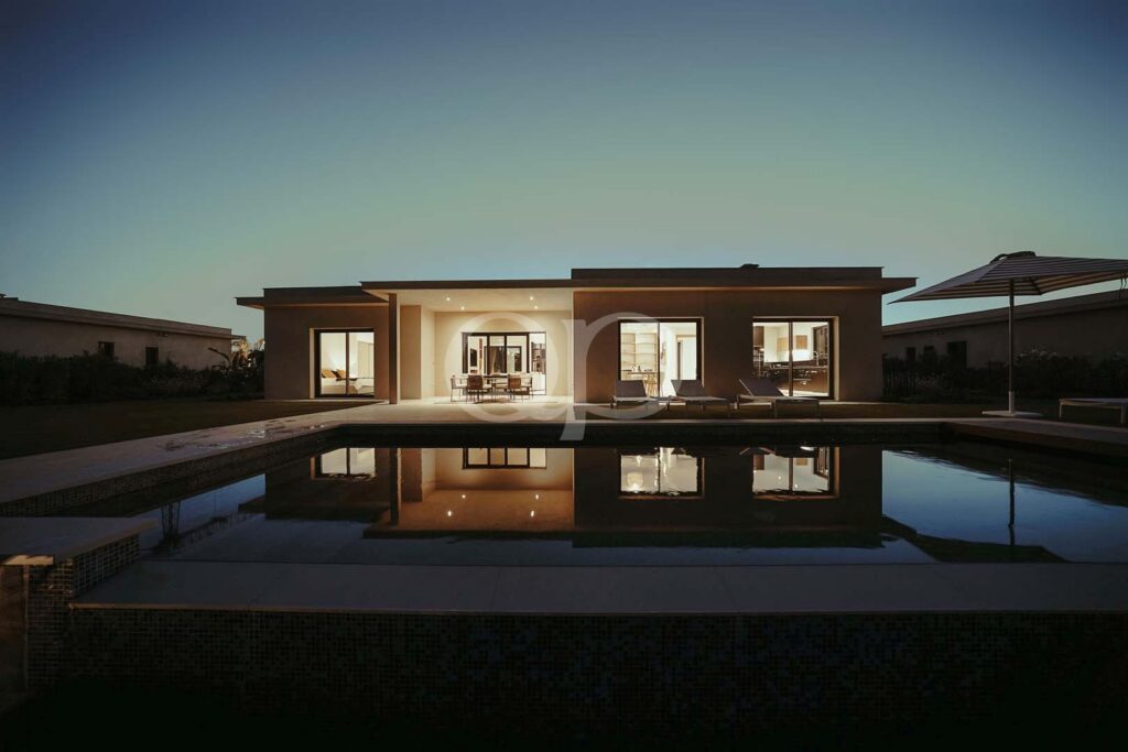 Casa moderna de um só piso com grandes portas de vidro, área de refeições ao ar livre e iluminação interior acolhedora, que se reflete na piscina ao anoitecer.