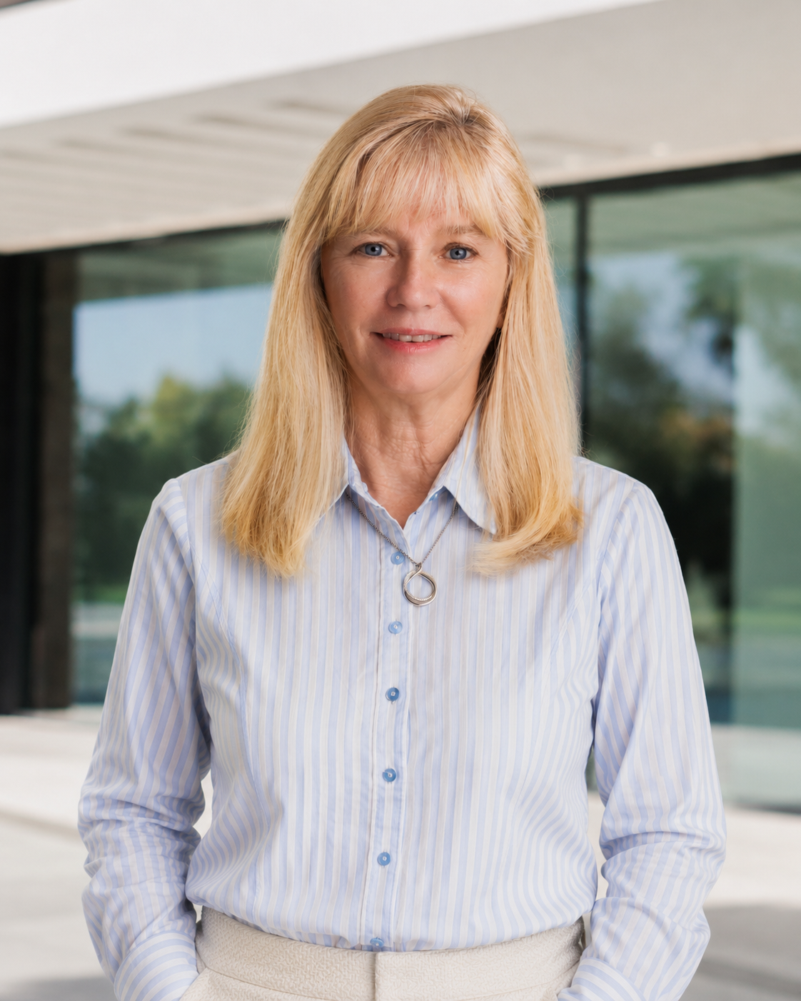 Portrait of a smiling blonde woman in a light blue striped shirt standing outdoors in front of a modern glass building