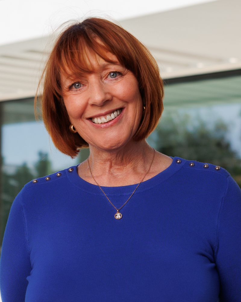 Smiling woman with short red hair in a blue sweater, gold necklace, outdoors.