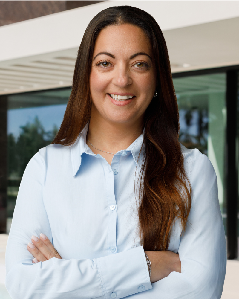 Mulher sorridente, com uma camisa de botões azul-clara e os braços cruzados, ao ar livre, junto a um edifício de escritórios moderno.