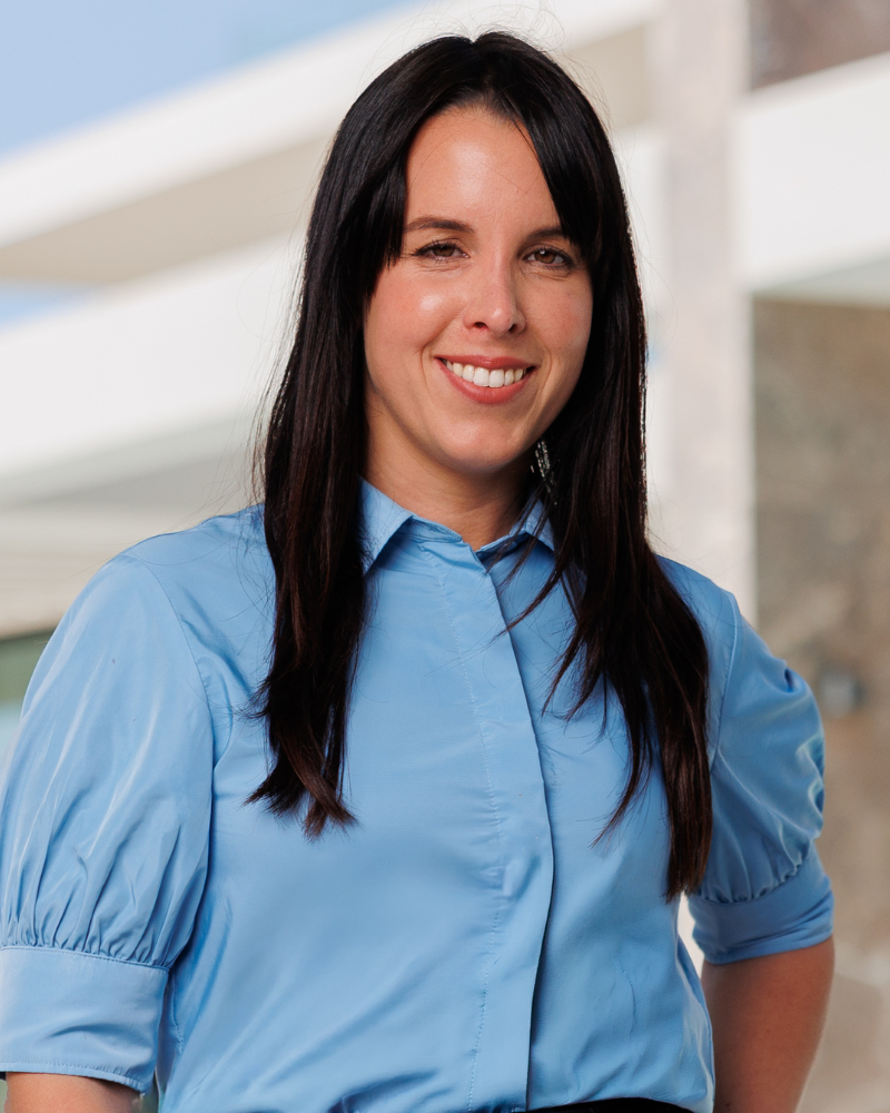 Mulher sorridente, com cabelo comprido e escuro, vestida com uma blusa azul-clara, ao ar livre, a posar para um retrato.