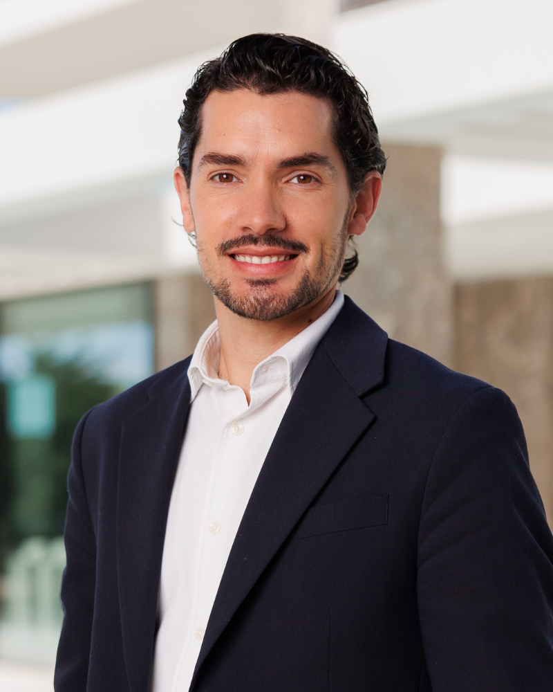 Photo de portrait professionnelle d'un homme vêtu d'un blazer bleu marine et d'une chemise blanche, souriant en extérieur près d'un immeuble moderne.