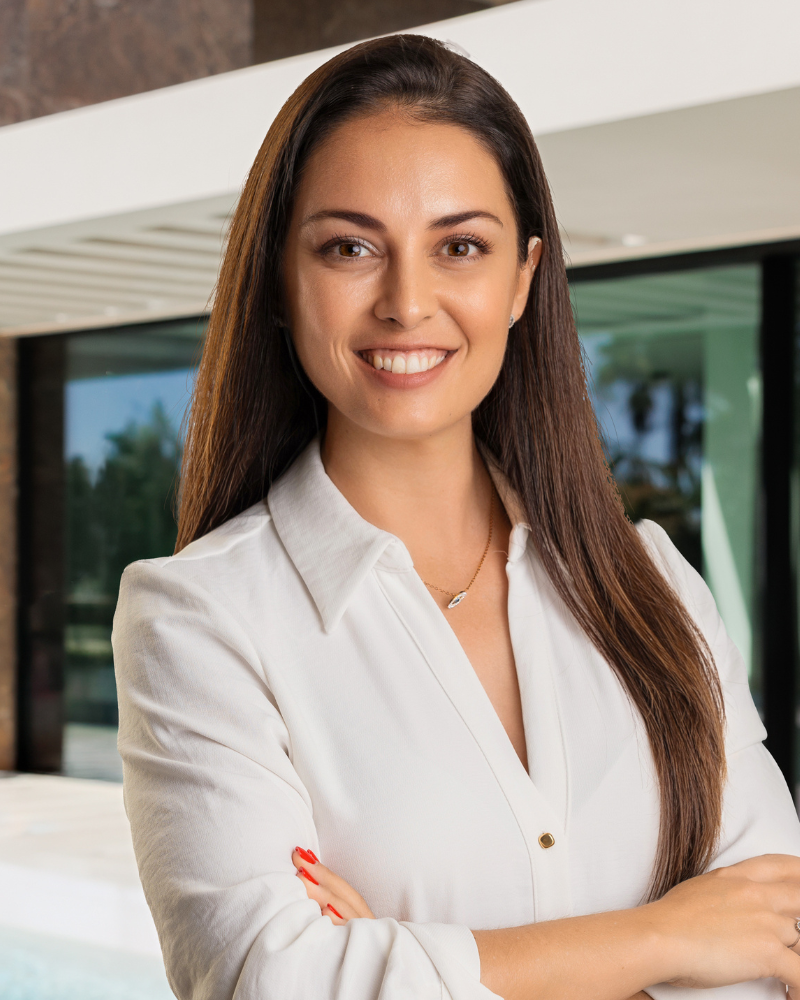 Femme d'affaires aux longs cheveux foncés, vêtue d'un chemisier blanc, souriante, les bras croisés, à l'extérieur près d'un immeuble moderne.