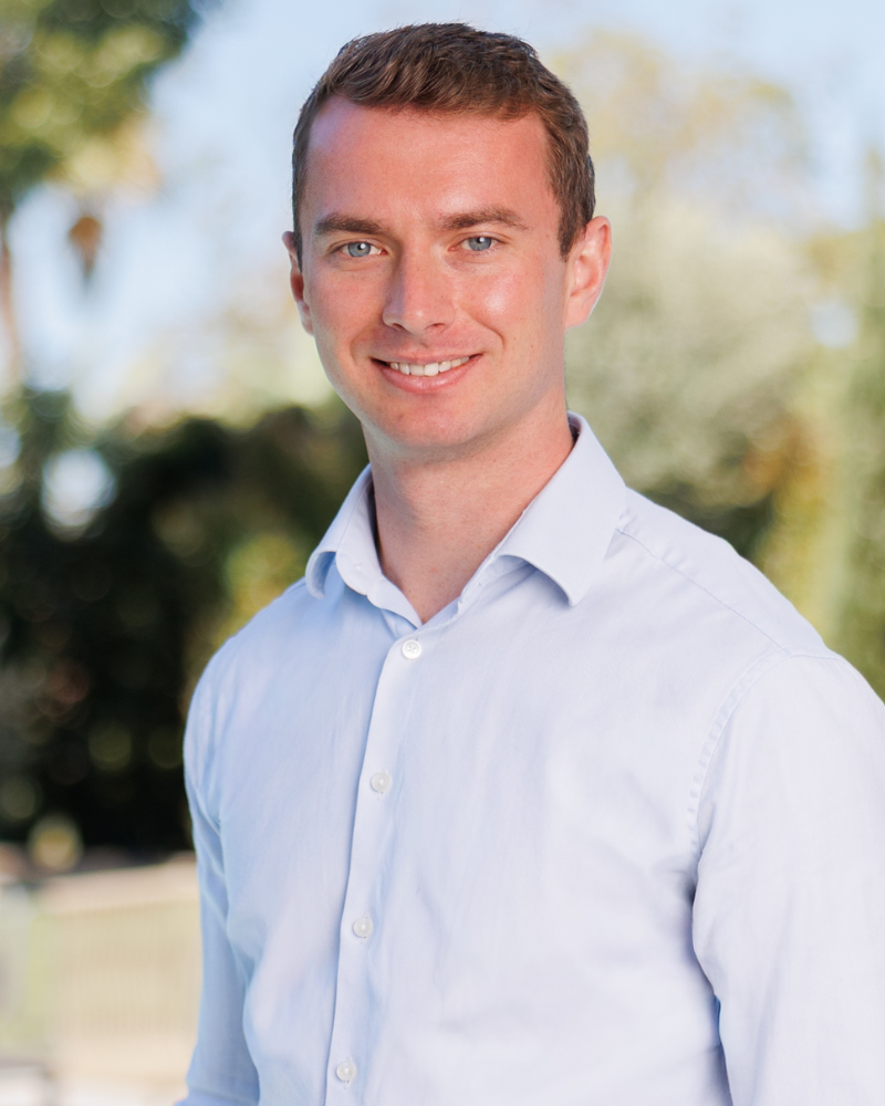Portrait of a smiling man in a light blue dress shirt outdoors, with blurred greenery in the background.