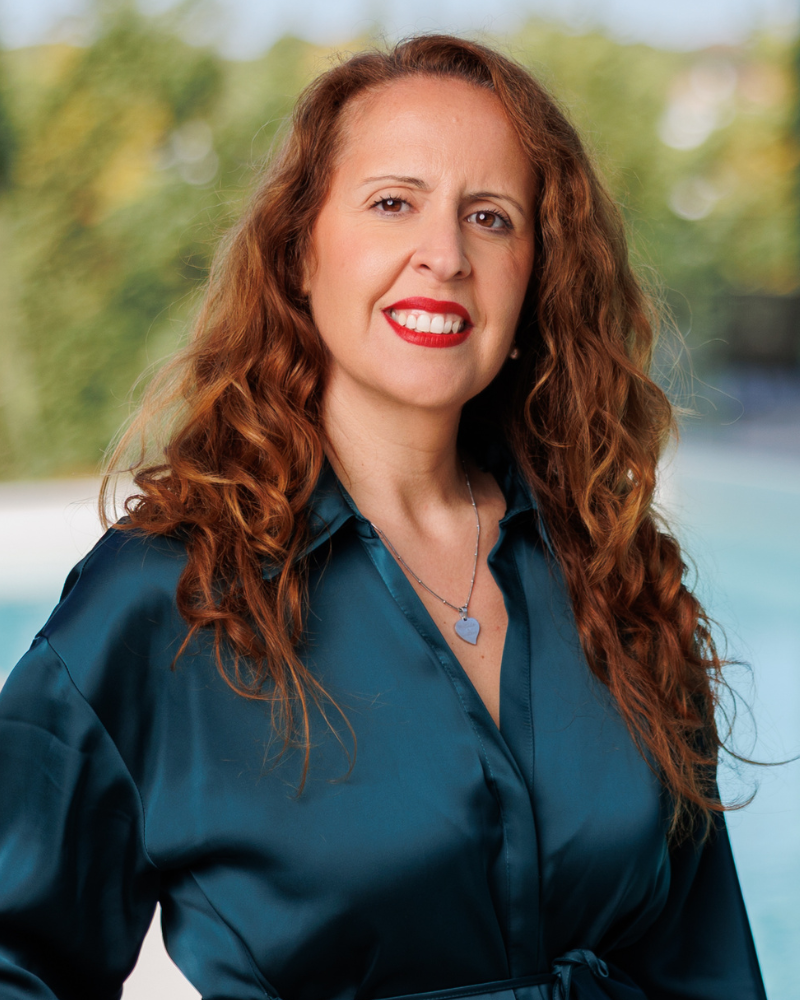 Portrait of a smiling woman with long curly red hair, wearing a teal satin blouse and a heart pendant necklace, outdoors near a pool.