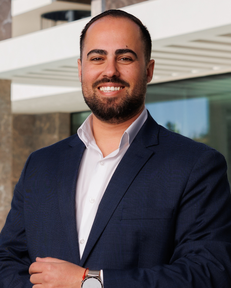 Smiling man in a navy suit and white shirt posing outdoors in front of a modern building.