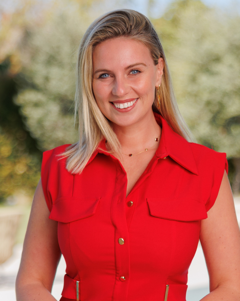 Portrait d'une femme blonde souriante vêtue d'un chemisier rouge sans manches, à l'extérieur, sur un fond vert flou.