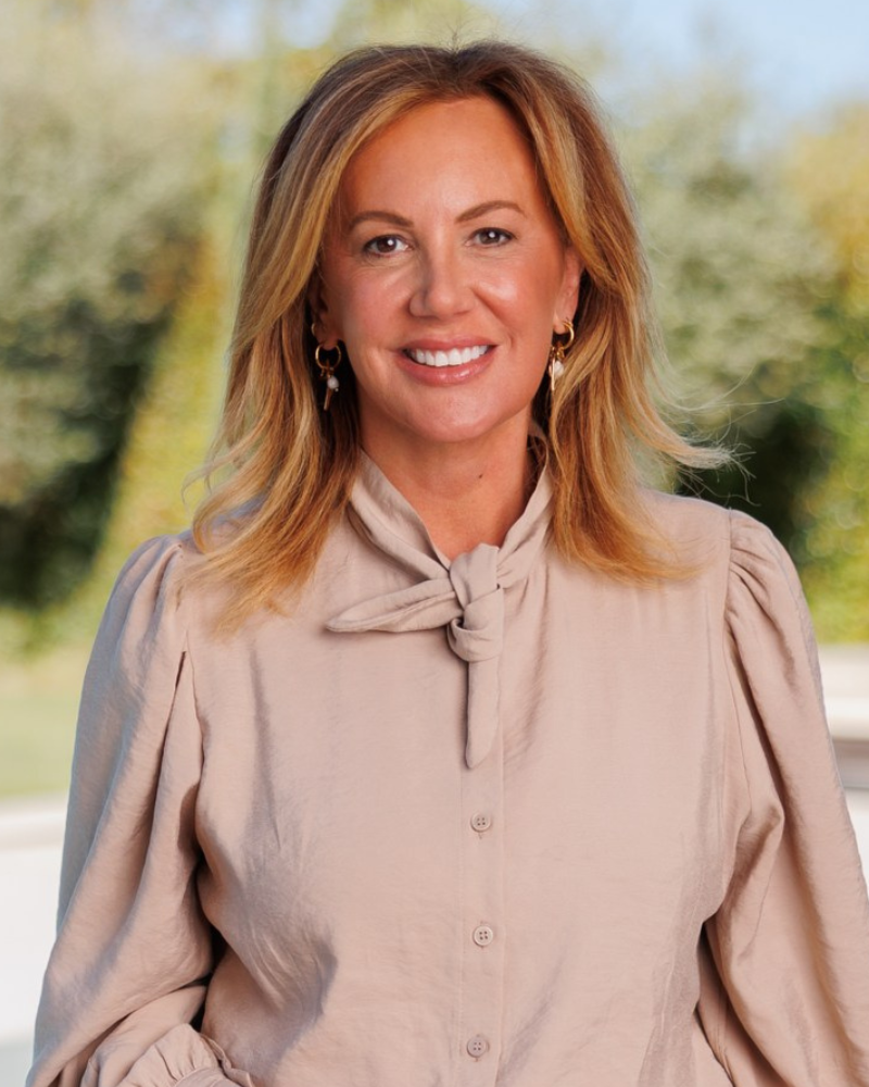 Portrait of a smiling woman with blonde hair wearing a beige blouse with a bow, outdoors with greenery in the background.