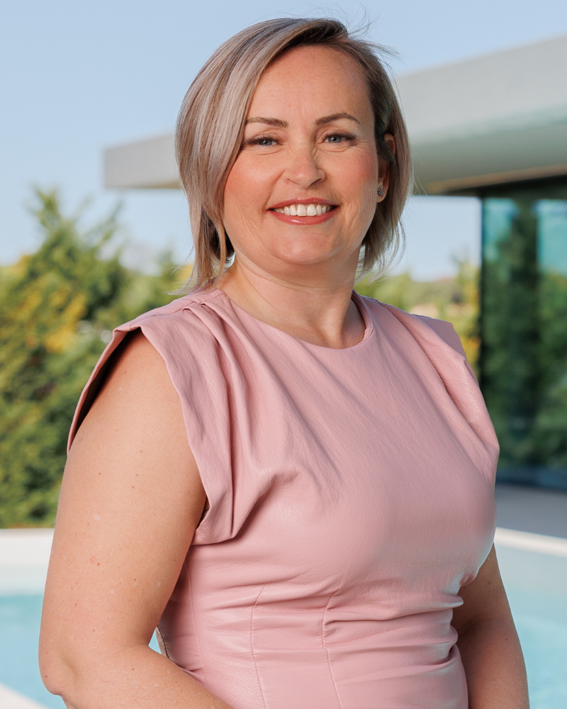 Portrait of a smiling woman in a pink sleeveless dress outdoors, with greenery and a modern building in the background.