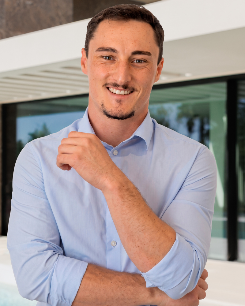 Smiling man in a light blue button-down shirt standing outdoors near a modern building.